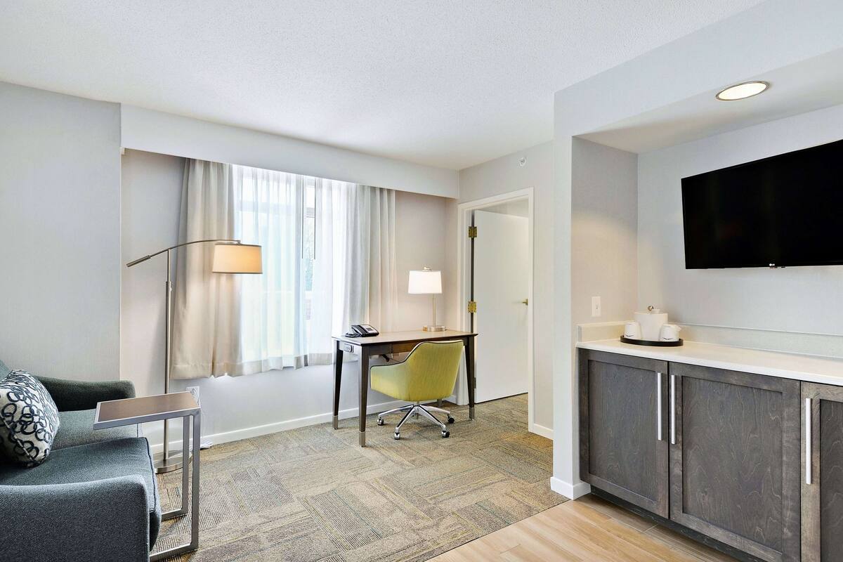 A hotel room with a sofa, desk, chair, and a flat-screen TV mounted on the wall.