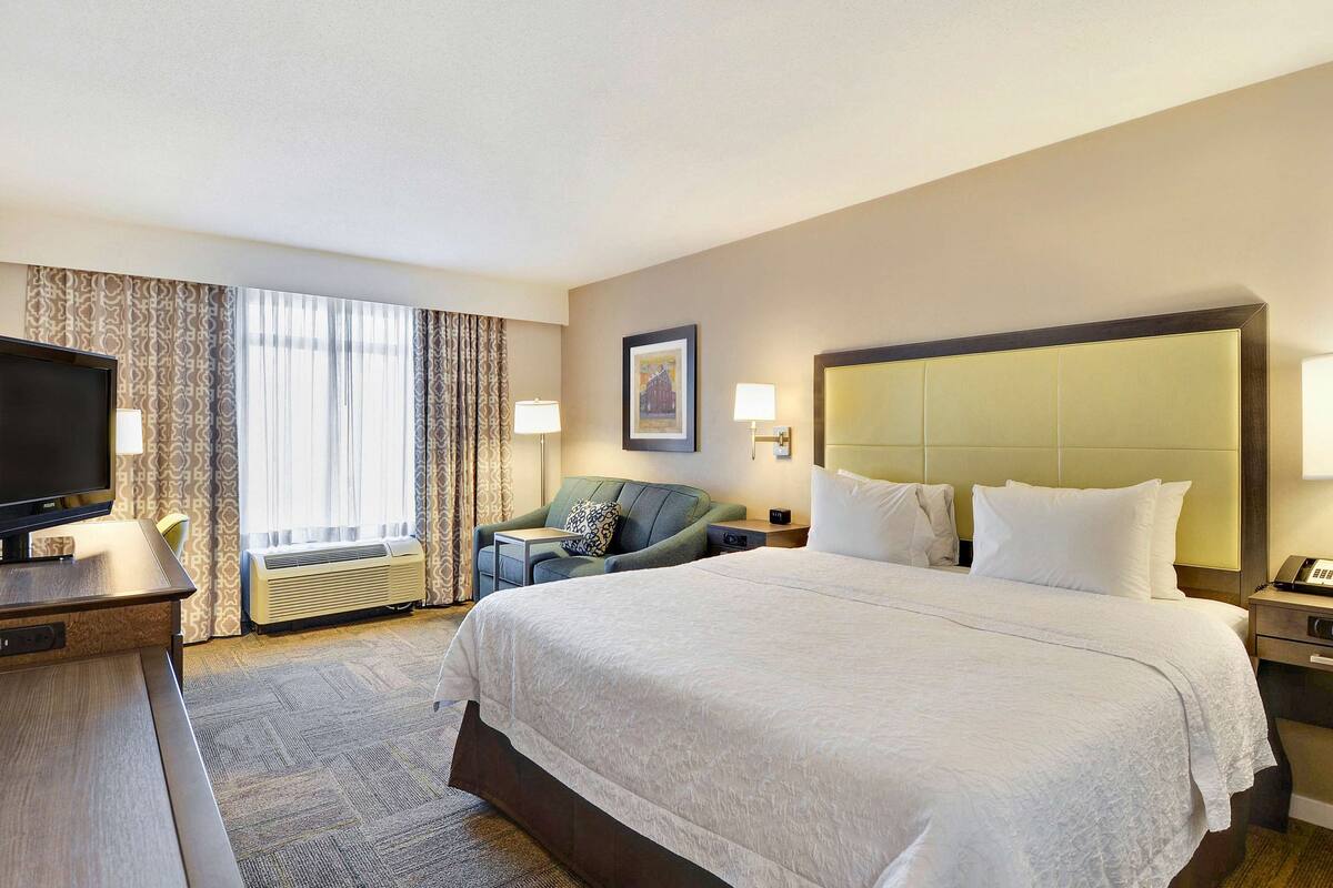 A hotel room with a large bed, a desk with a television, a chair, and a window with curtains.
