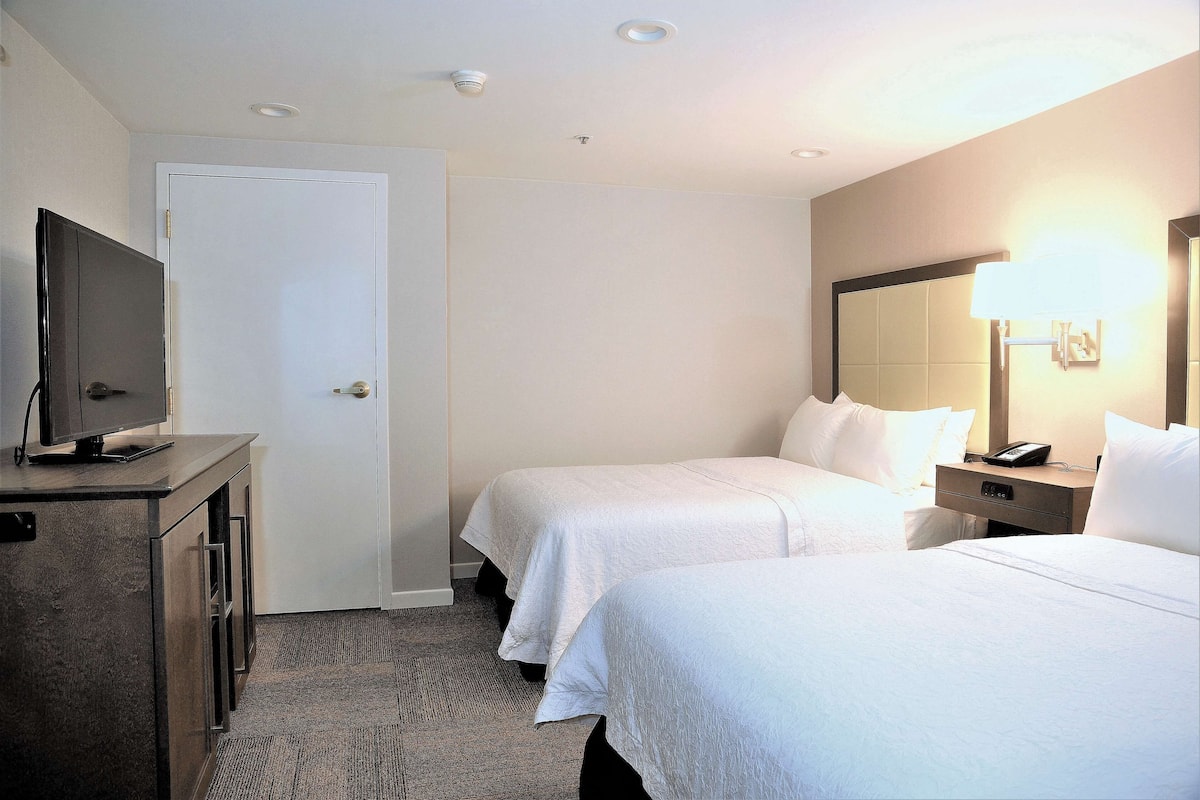 A hotel room with two beds, a desk with a TV, a bedside table with a lamp, and a door.