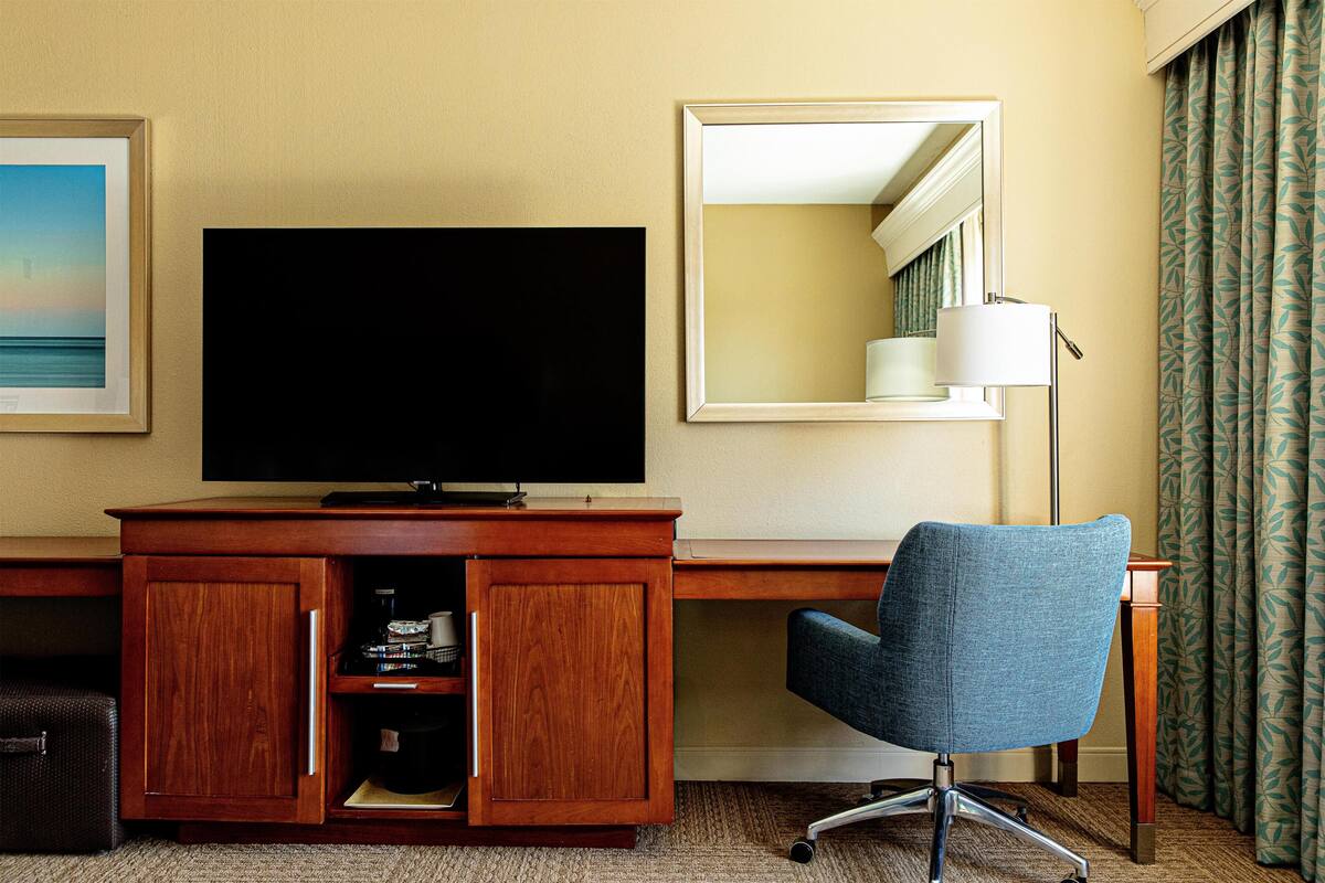 A hotel room with a wooden desk, a television, a blue chair, a mirror, and a patterned curtain.