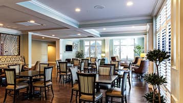 A spacious dining area with multiple tables and chairs, a television mounted on the wall, and large windows allowing natural light.