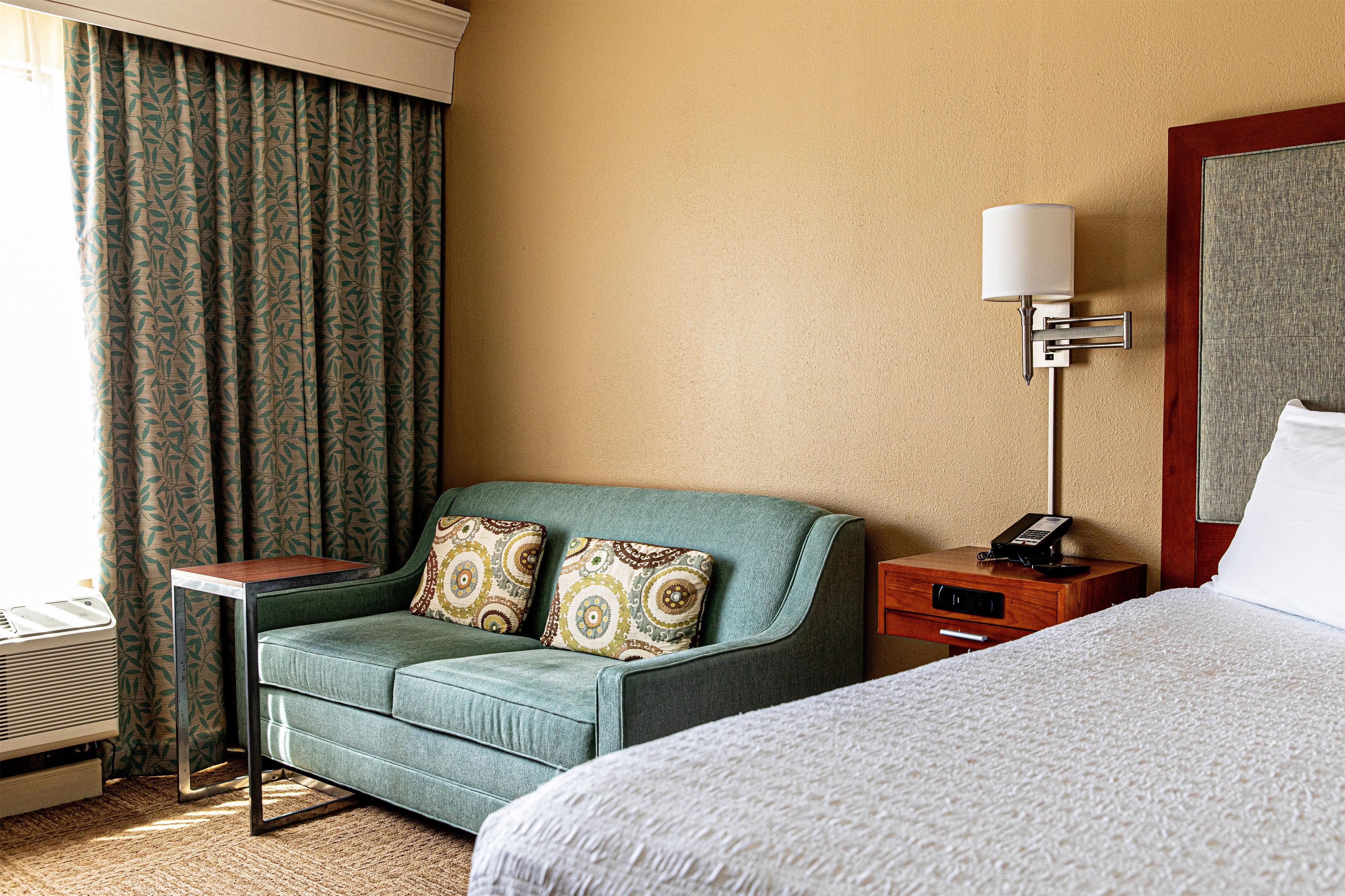 A hotel room with a bed, a green armchair with patterned cushions, a bedside table with a lamp, and a wall-mounted telephone.