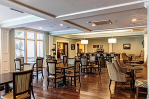 Uma ampla área de refeições com várias mesas e cadeiras, um bar ao fundo e grandes janelas com persianas.