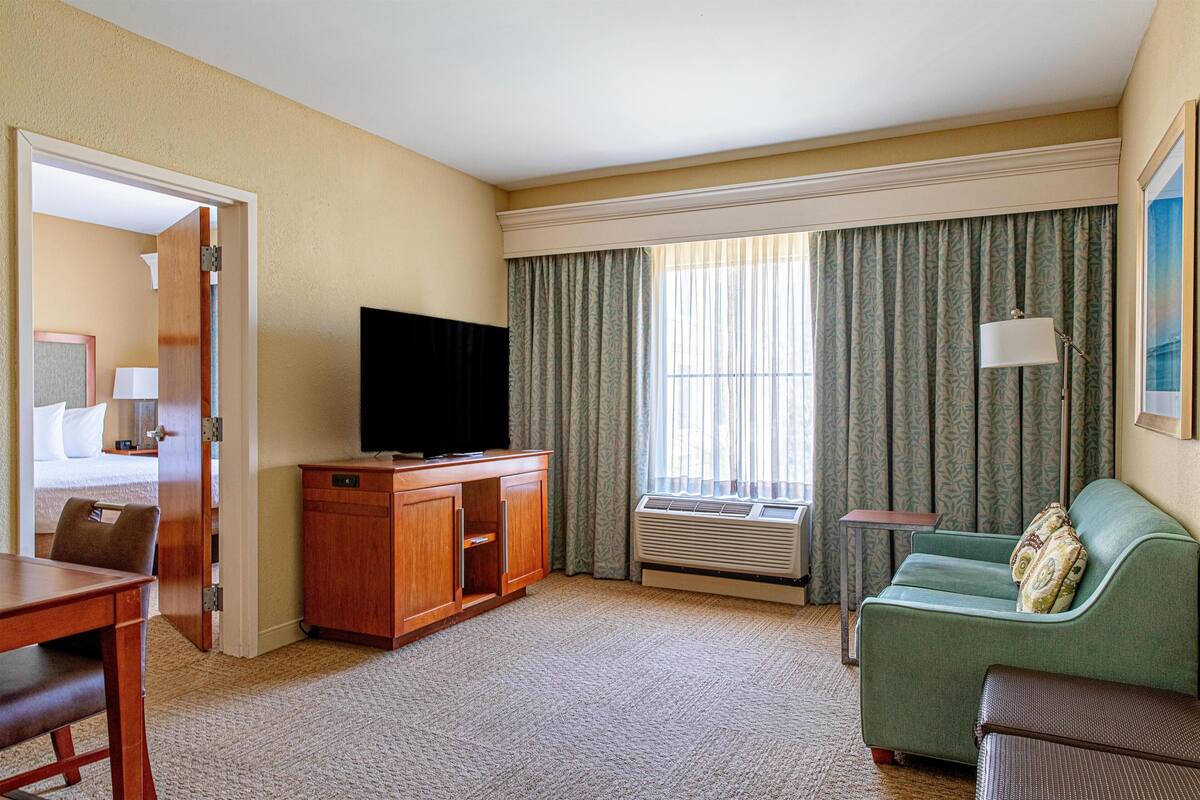 A hotel room with a television, a sofa, a dining table, and a window with curtains.