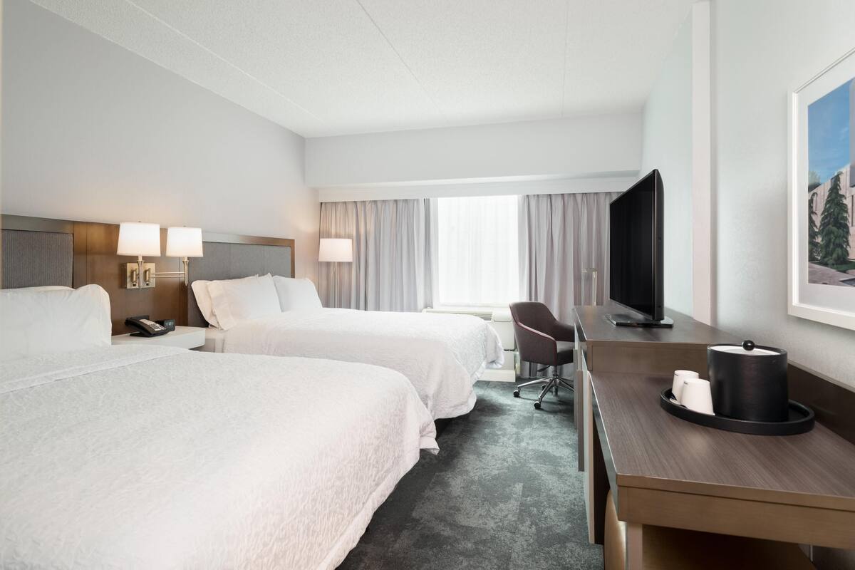 A hotel room with two beds, a desk, a chair, a television, and a painting on the wall.