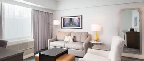 A hotel room with a sofa, armchair, coffee table, side table with lamp, and a large mirror.