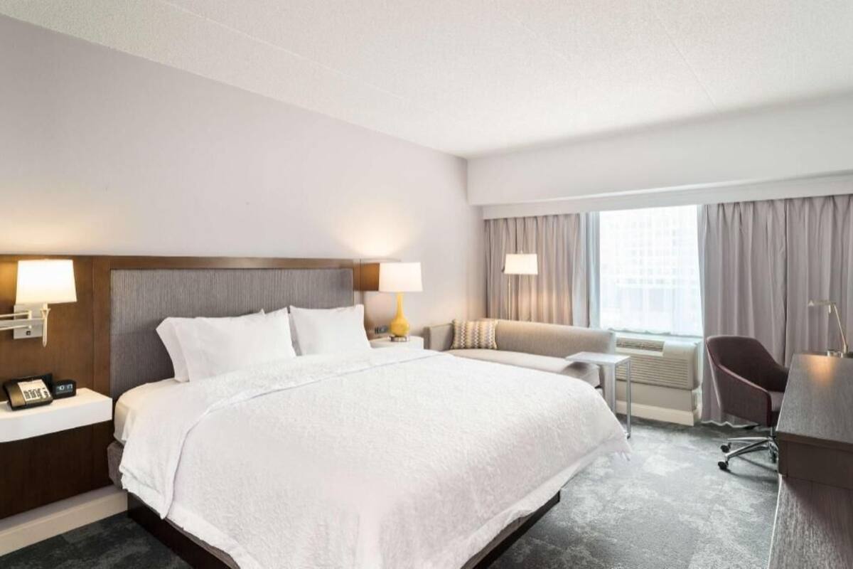 A hotel room with a large bed, bedside tables with lamps, a sofa, a desk, and a chair.