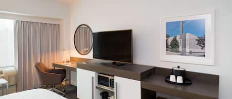 A hotel room with a bed, a desk with a flat-screen TV, a chair, a microwave, a coffee maker, and a framed picture on the wall.