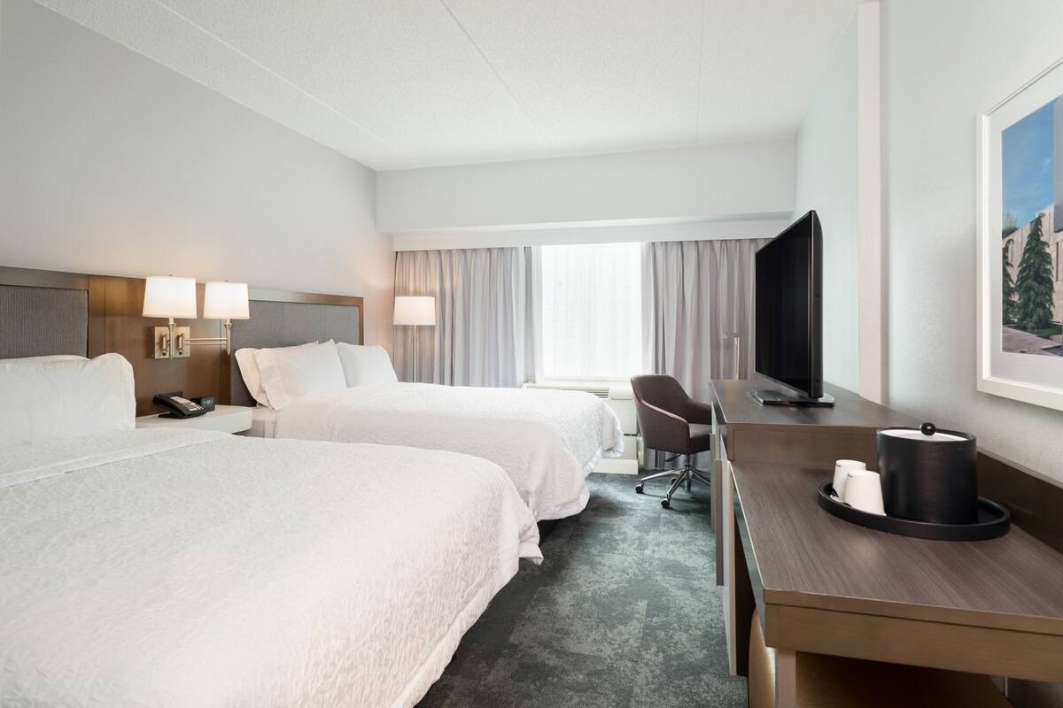 A hotel room with two beds, a desk, a chair, a television, and a painting on the wall.