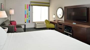 A hotel room with a bed, a desk with a television, a chair, a lamp, a mirror, a window, and a wall with colorful decor.