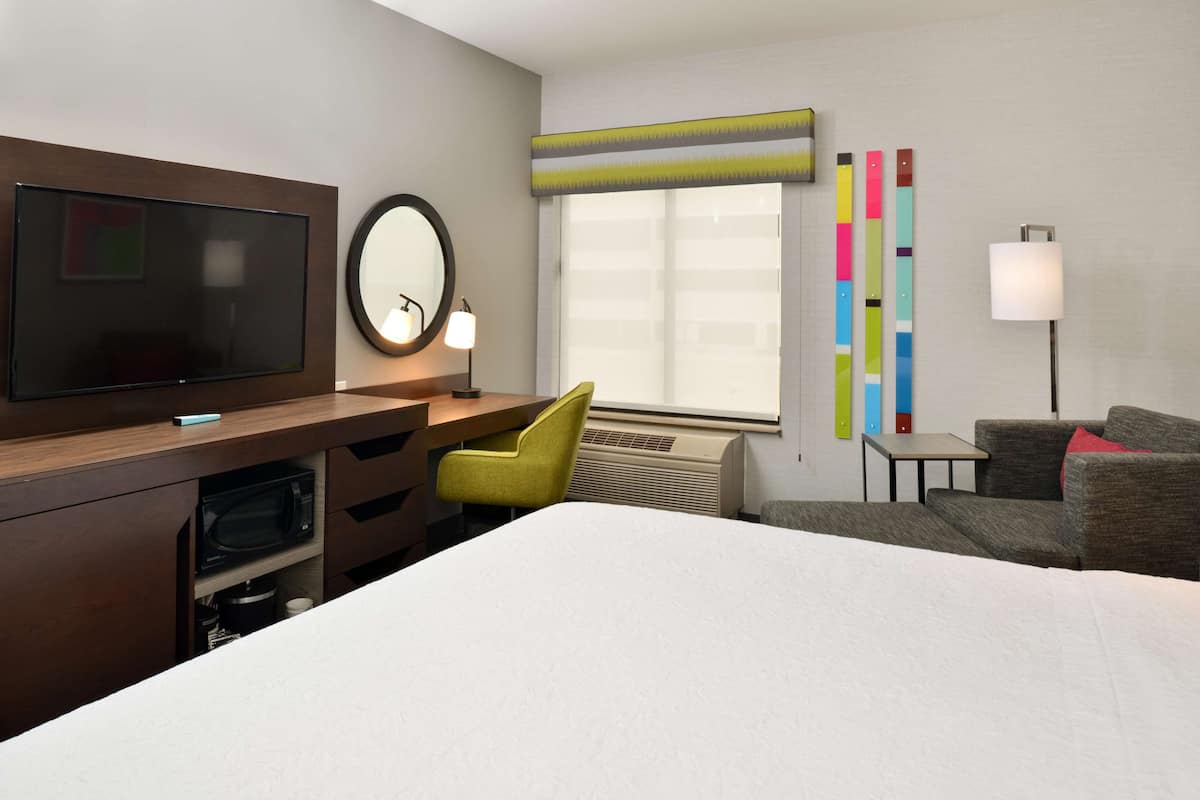 Habitación de hotel con cama, escritorio con televisor de pantalla plana, silla, mesita, lámpara y ventana con persianas coloridas.