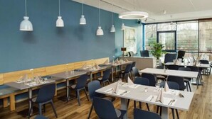 A modern dining area with wooden tables and blue chairs, featuring pendant lighting and large windows. - Hotel INNdependence (Mainz)