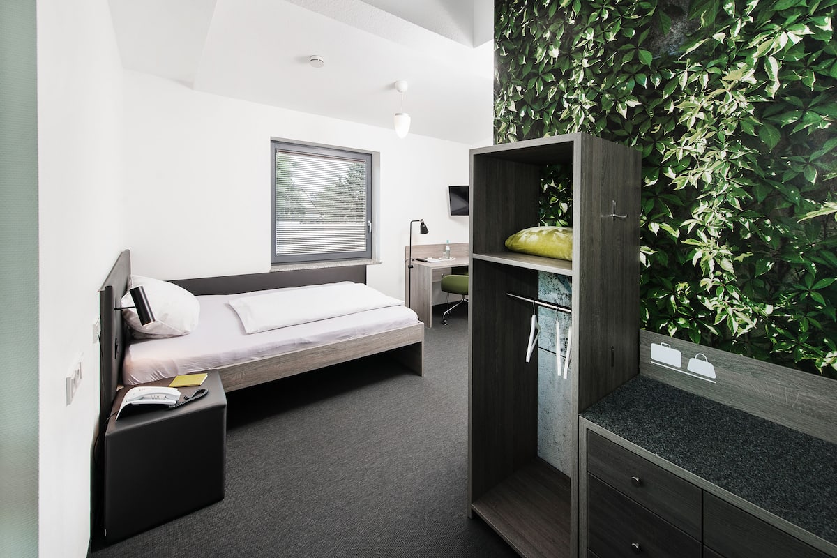 A modern hotel room with a bed, a desk, a wardrobe, and a green leafy wall.