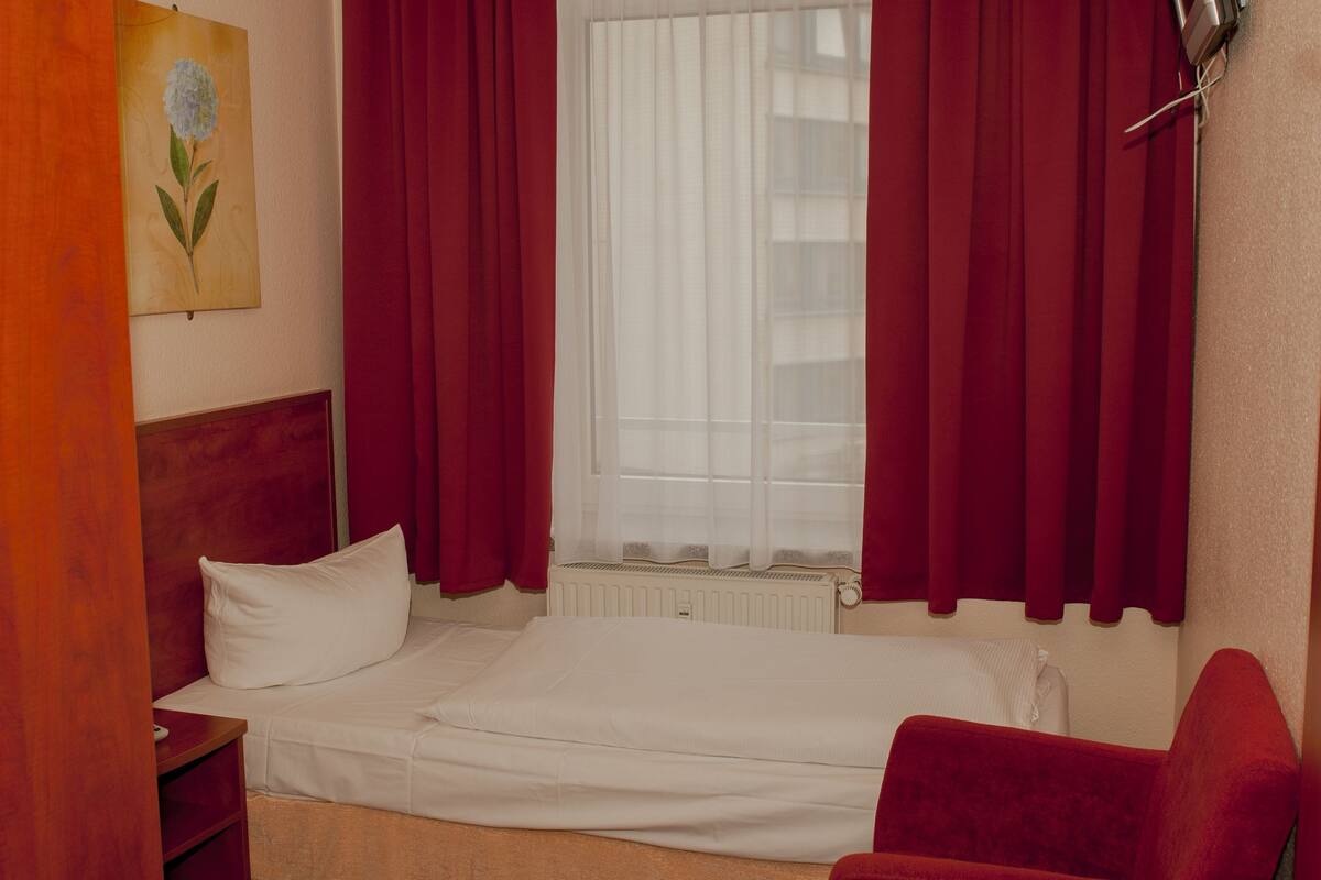 Ein Hotelzimmer mit Bett, einem roten Sessel, einem Bild an der Wand und einem Fenster mit Vorhängen.