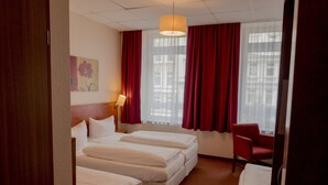 Une chambre d’hôtel avec trois lits, une chaise rouge, un bureau et une fenêtre avec des rideaux rouges.