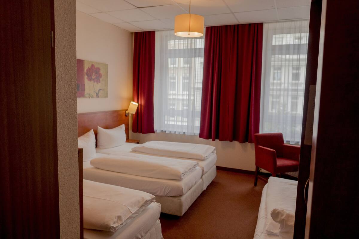 Ein Hotelzimmer mit drei Betten, einem roten Sessel, einem Schreibtisch und einem Fenster mit roten Vorhängen.