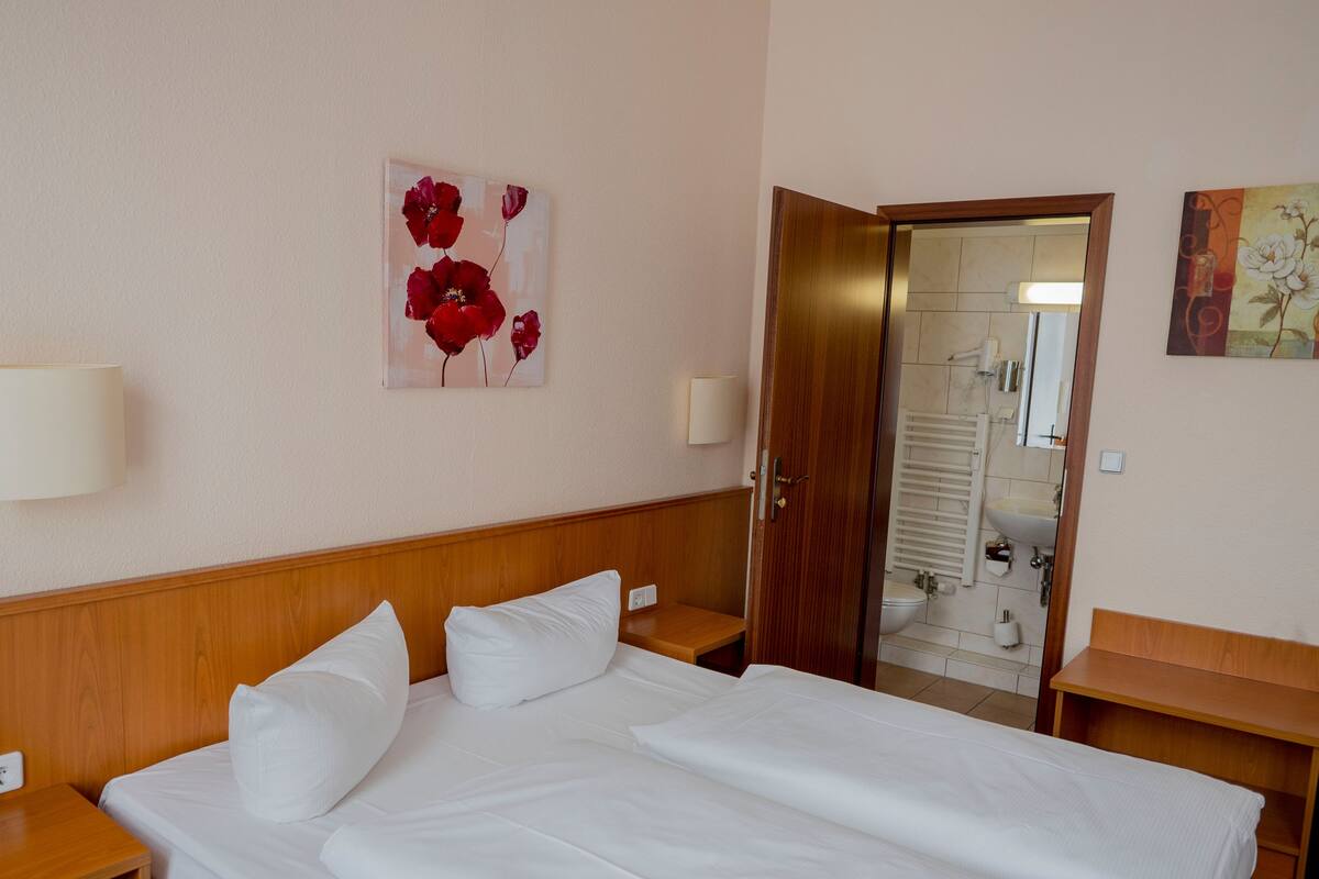 Ein Hotelzimmer mit einem hölzernen Kopfende, einem großen Bett mit weißer Bettwäsche, einem Nachttisch, einer Nachttischlampe, einem Gemälde von roten Blumen an der Wand und einem durchgegangenen Tür erkennbaren Badezimmer.
