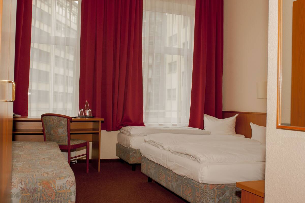Ein Hotelzimmer mit zwei Betten, einem Schreibtisch, einem Stuhl und einem Fenster mit roten Vorhängen.