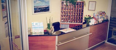 A reception desk with a wooden panel design, a framed cityscape picture, a plant, and a display of brochures.