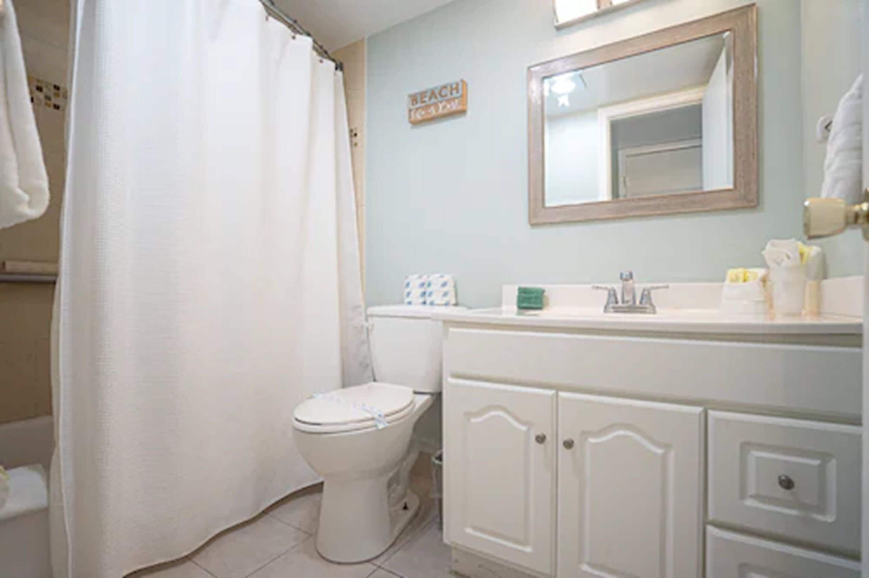 A bathroom with a white toilet, a shower curtain, a sink with toiletries, and a mirror above the sink.