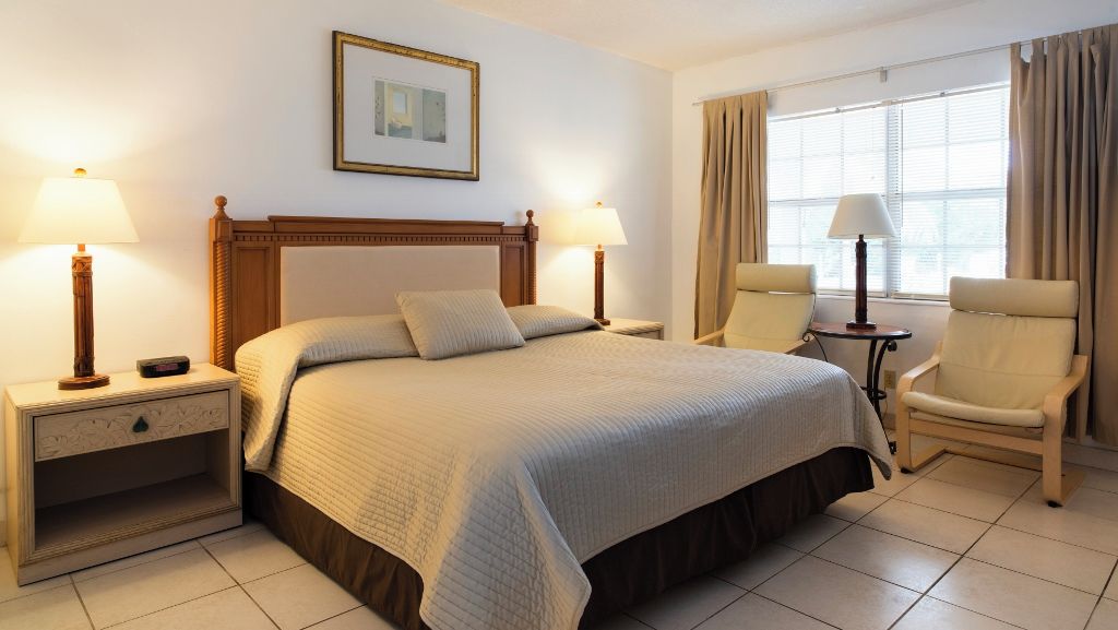 A hotel room with a bed, two bedside tables, a chair, a window with curtains, and a framed picture on the wall.
