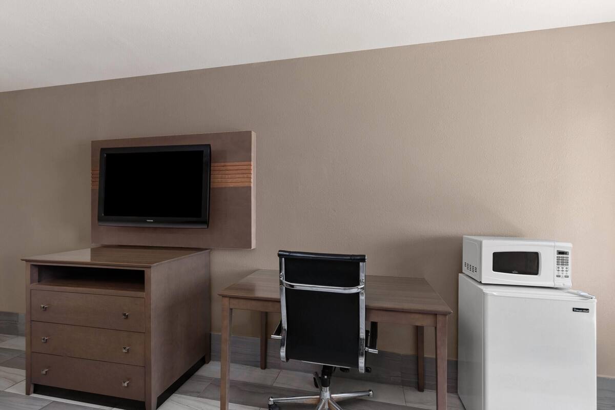A hotel room with a flat-screen TV mounted on the wall, a wooden desk, a black office chair, a built-in wooden cabinet, and a white refrigerator.