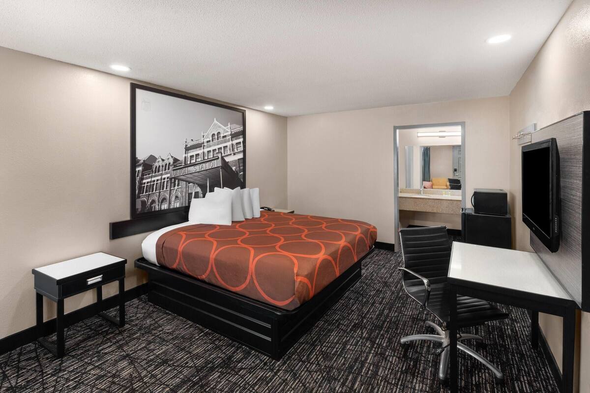 A hotel room with a bed, a desk, a chair, a television, a mirror, and a framed picture on the wall.