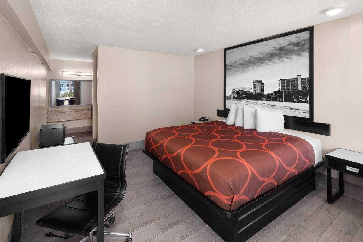 A hotel room with a bed, desk, chair, and a cityscape painting on the wall.