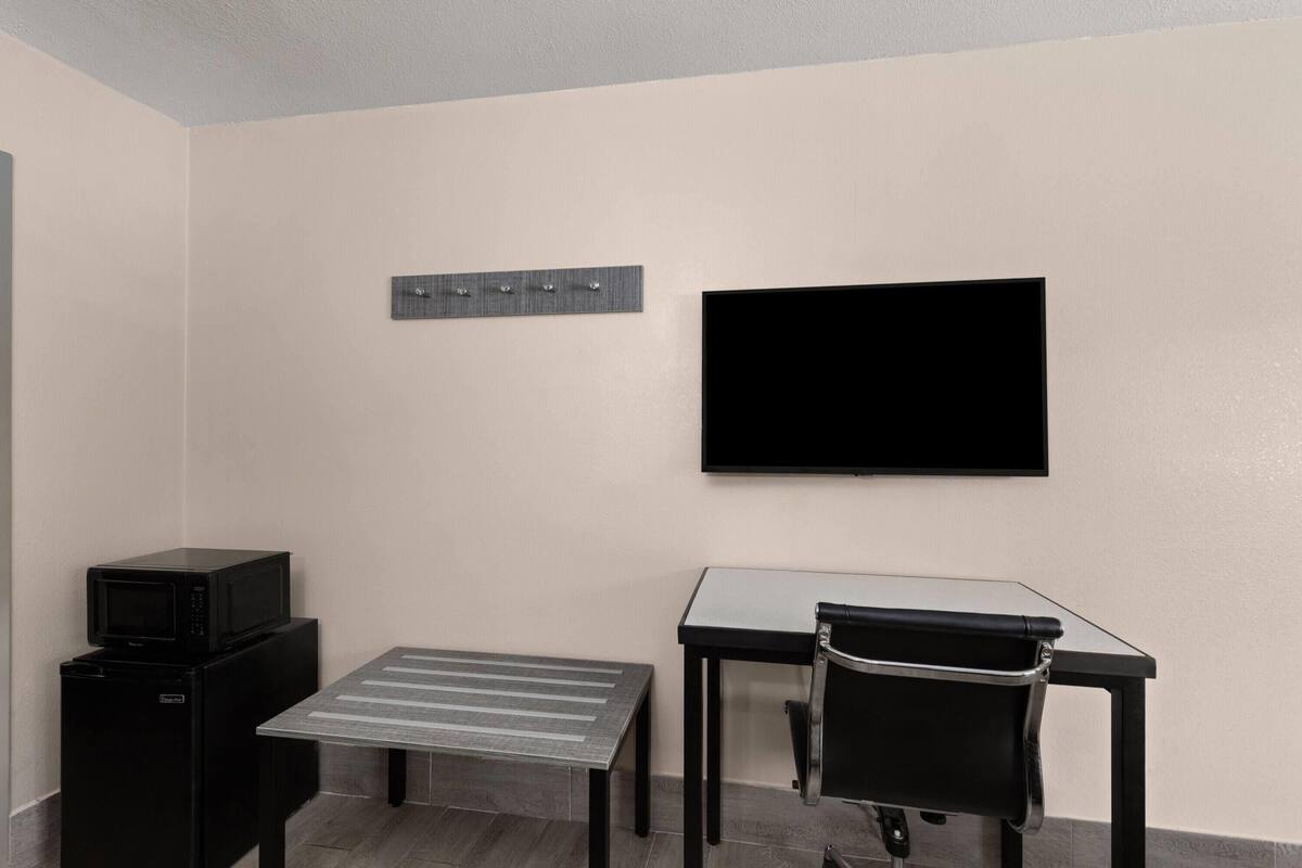 A small room with a flat-screen TV mounted on the wall, a small black refrigerator, a table with a chair, and a bench.