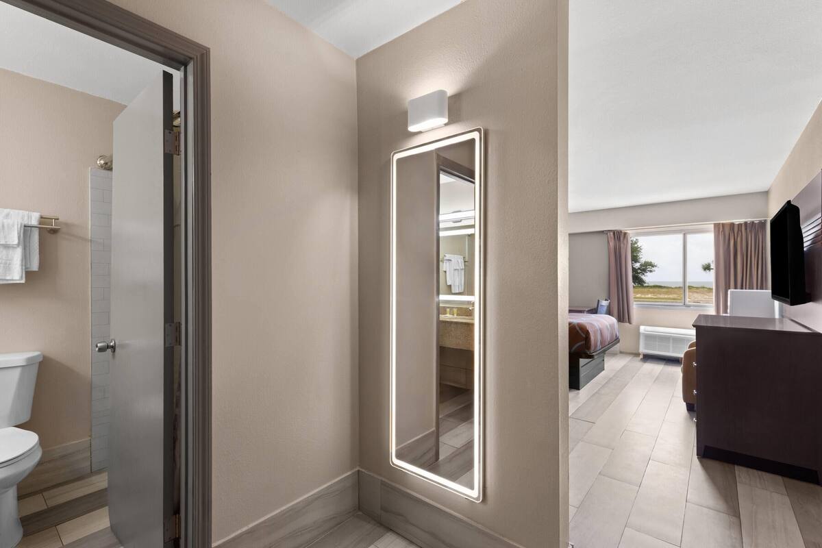 A hotel room with a bathroom, a bed, a desk, and a large mirror with lights.