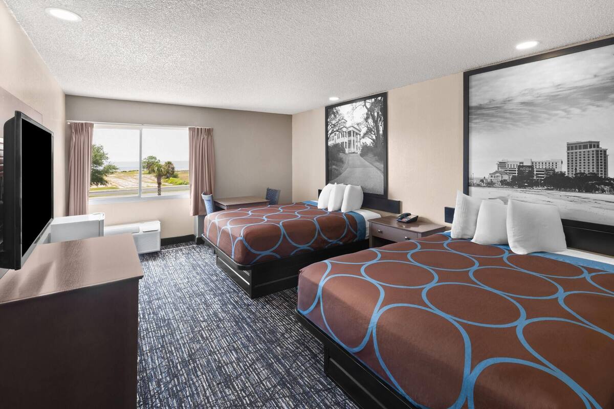 A hotel room with two beds, a desk, a chair, a TV, and a large window with curtains.
