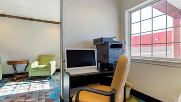 A hotel room with a desk, a computer, a printer, a window with a view of a red building, a patterned carpet, and a green armchair.