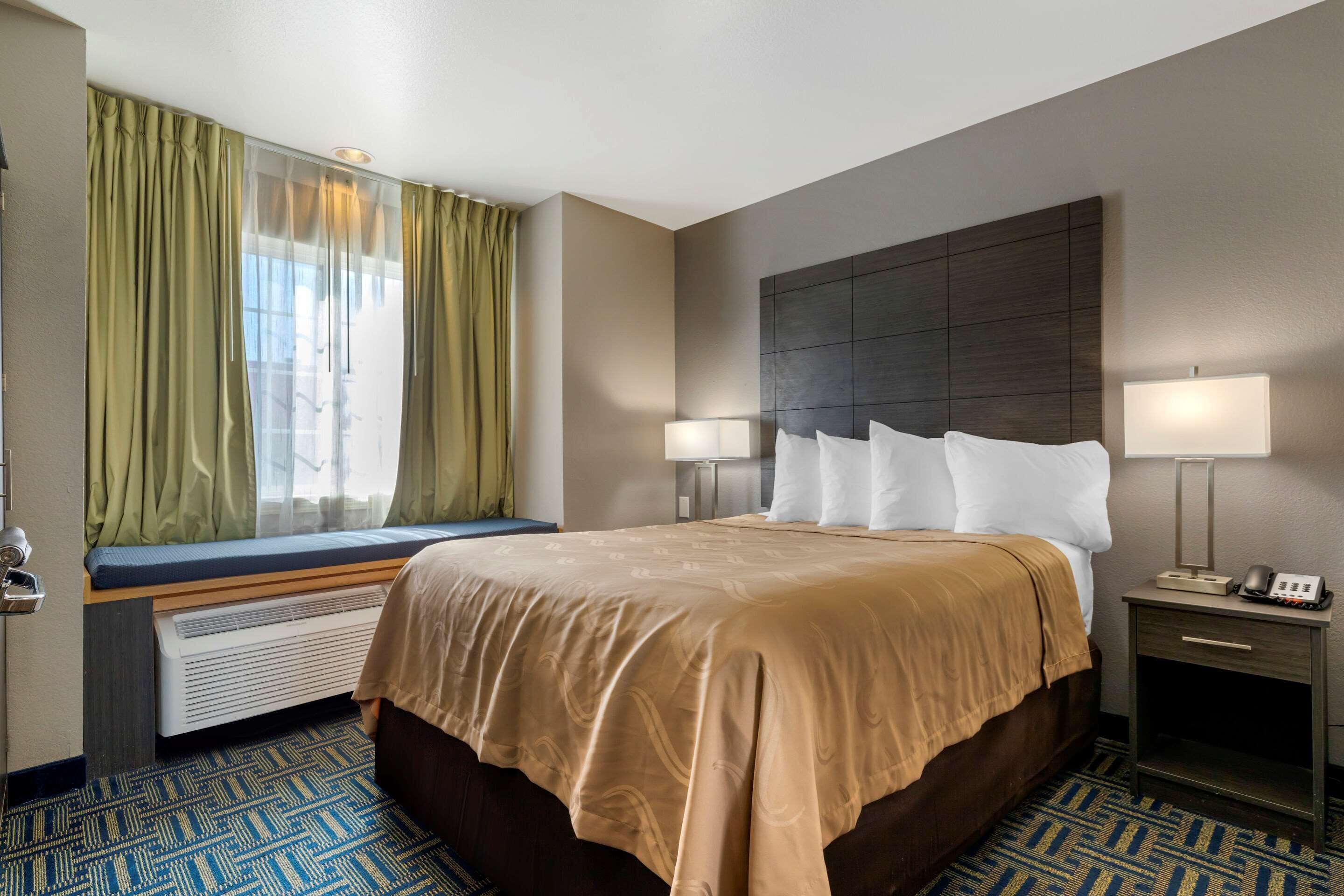 Habitación de hotel con una cama grande, una mesita de noche con lámpara, una ventana con cortinas y un banco empotrado debajo de la ventana.