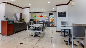 A hotel dining area with tables and chairs, a kitchen area with a refrigerator and coffee machine, and a sign indicating breakfast hours. - Quality Inn Sallisaw (Sallisaw)
