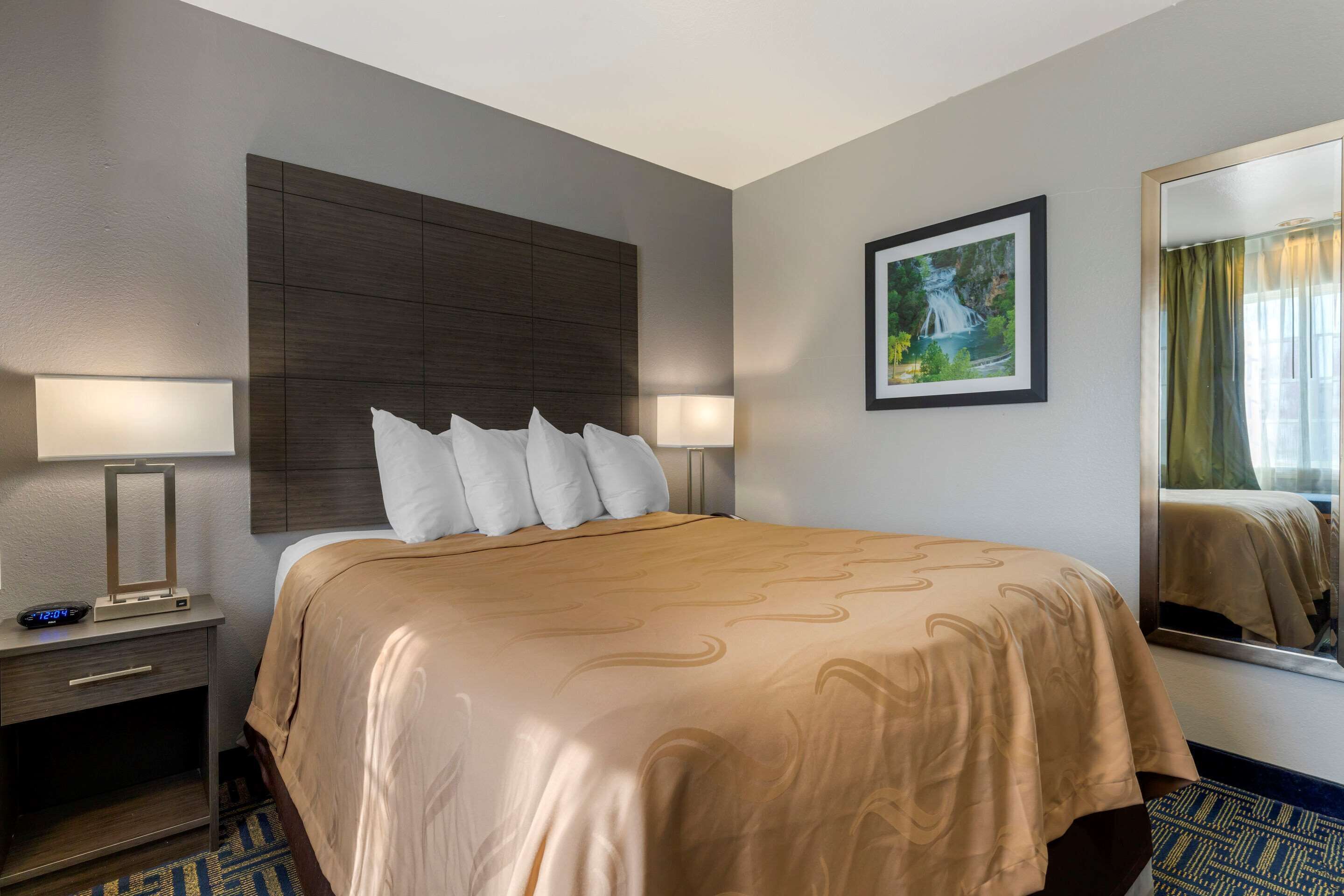 A hotel room with a large bed, two bedside tables with lamps, a framed picture on the wall, and a mirror reflecting part of the room.
