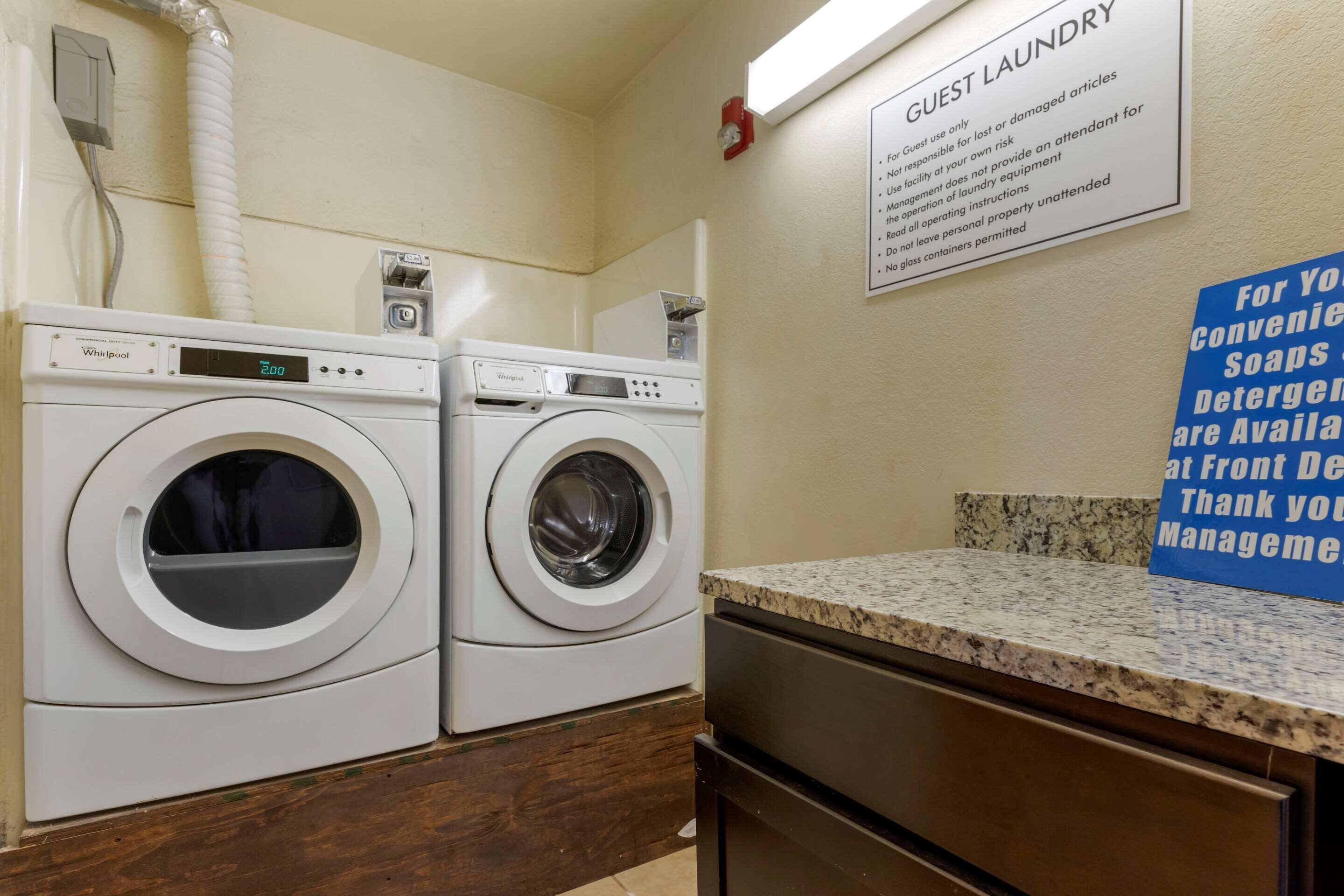 A guest laundry area with two Whirlpool washers and dryers, a sign detailing laundry policies, and a countertop with a sign about available soaps and detergents.