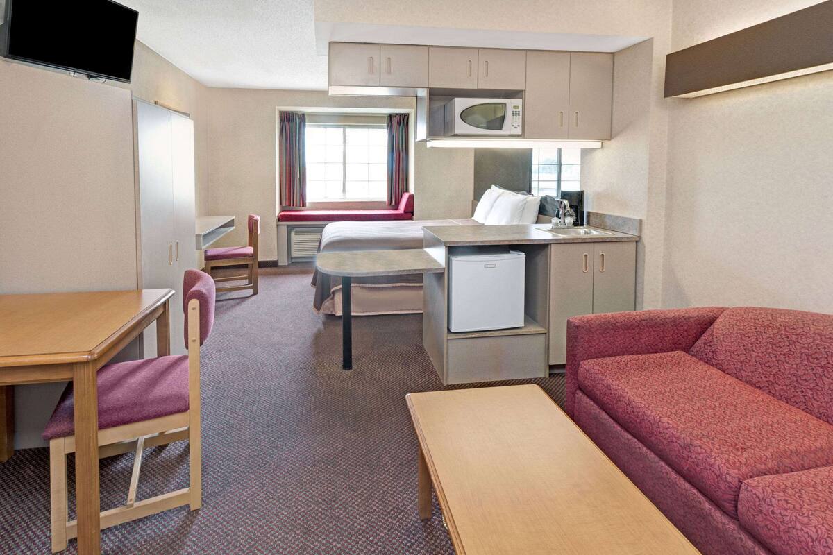 A compact hotel room with a kitchenette, a dining area, a sofa, and a TV.