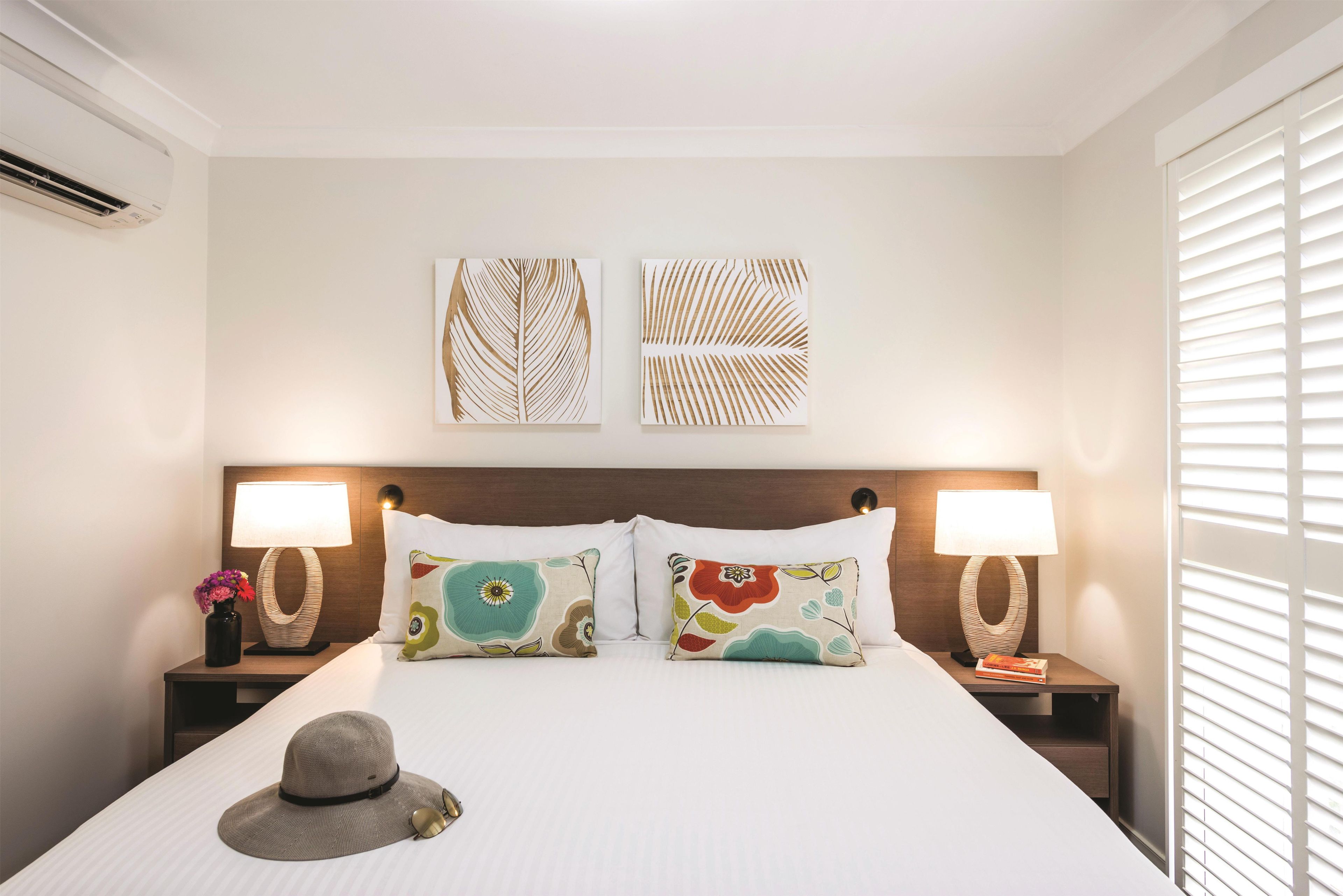 Una camera d'albergo con un letto, due comodini, due lampade a parete, un cappello decorativo sul letto e due opere d'arte a parete.