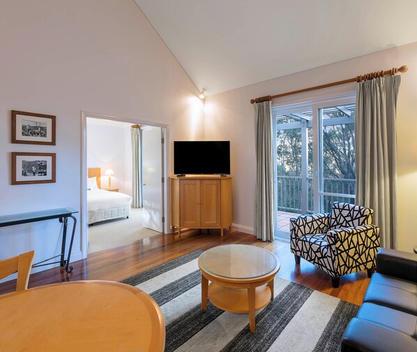 Una habitación de hotel moderna con una cama grande, un televisor sobre un mueble de madera, una mesa de vidrio, un sillón estampado y un sofá.