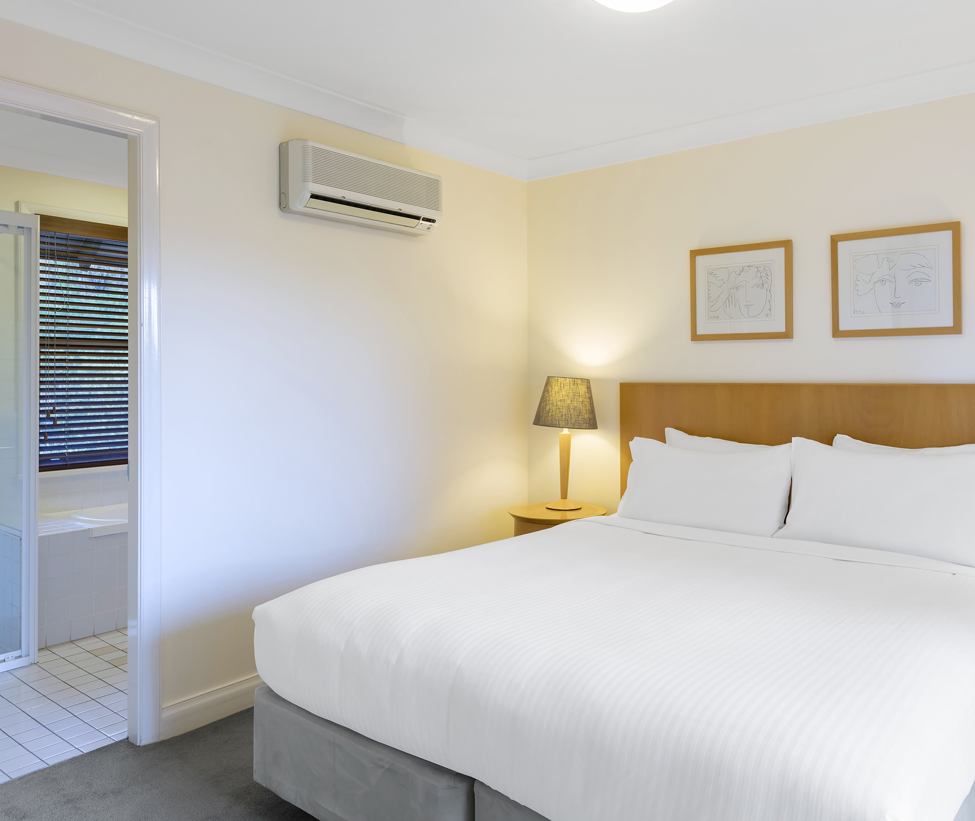 A hotel room with a bed, a nightstand, a lamp, an air conditioning unit, and two framed pictures on the wall.