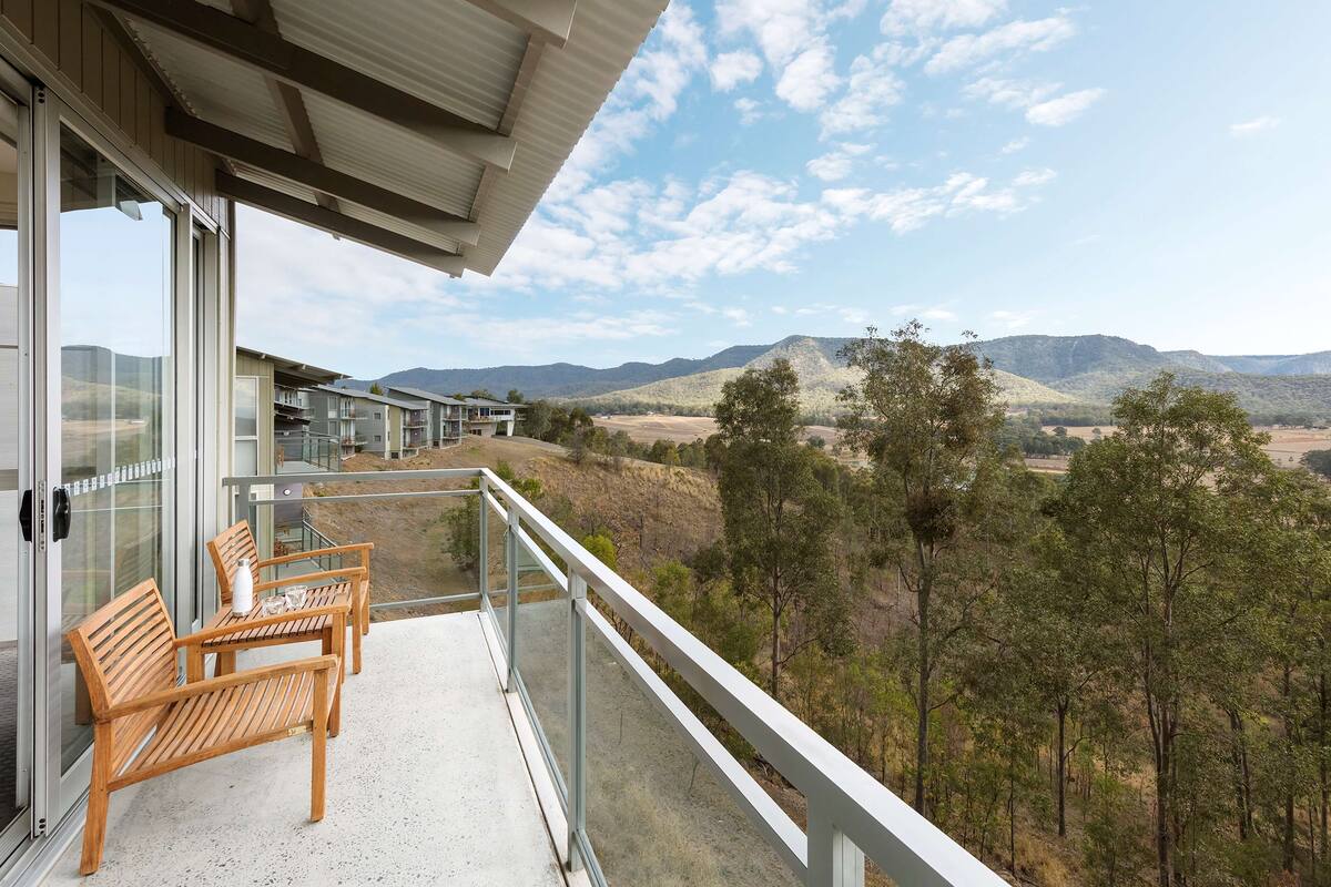 Un balcon doté de mobilier en bois et d’une balustrade en verre, offrant une vue sur un paysage montagneux.