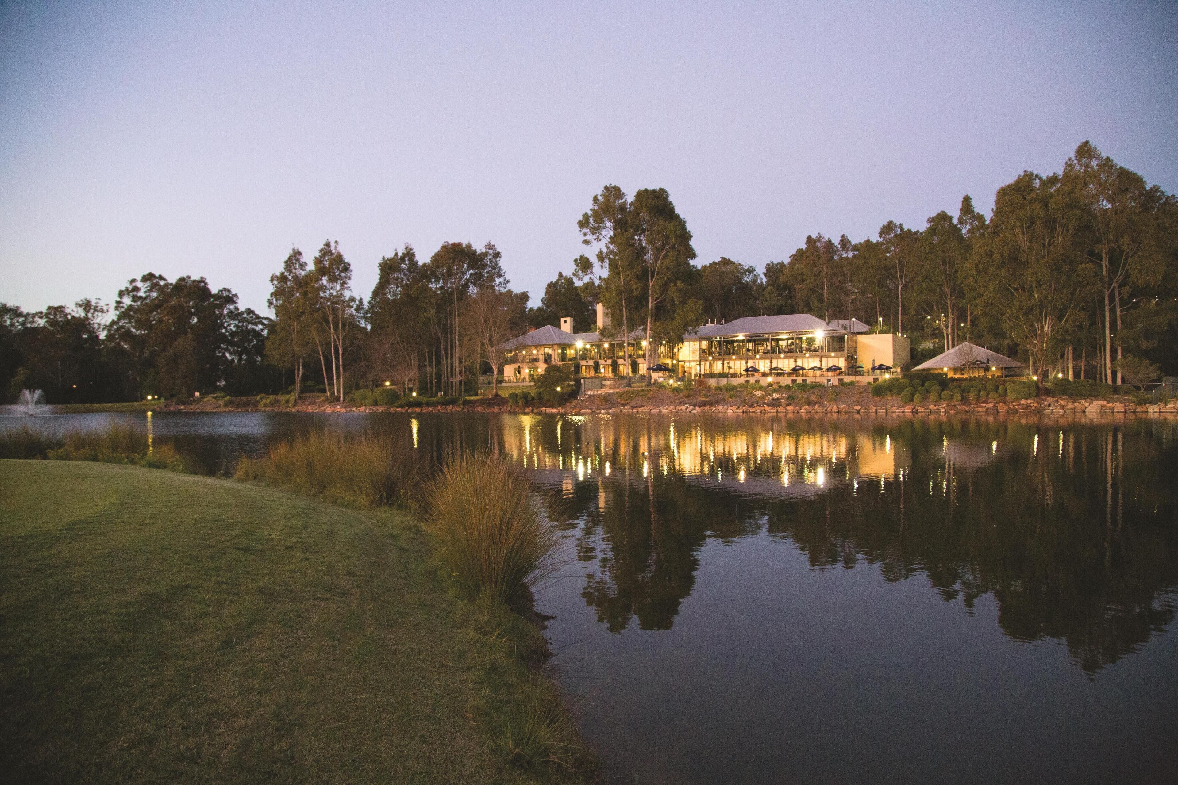 Hotel frente a un lago, con su reflejo en el agua, rodeado de árboles y un césped cuidado.