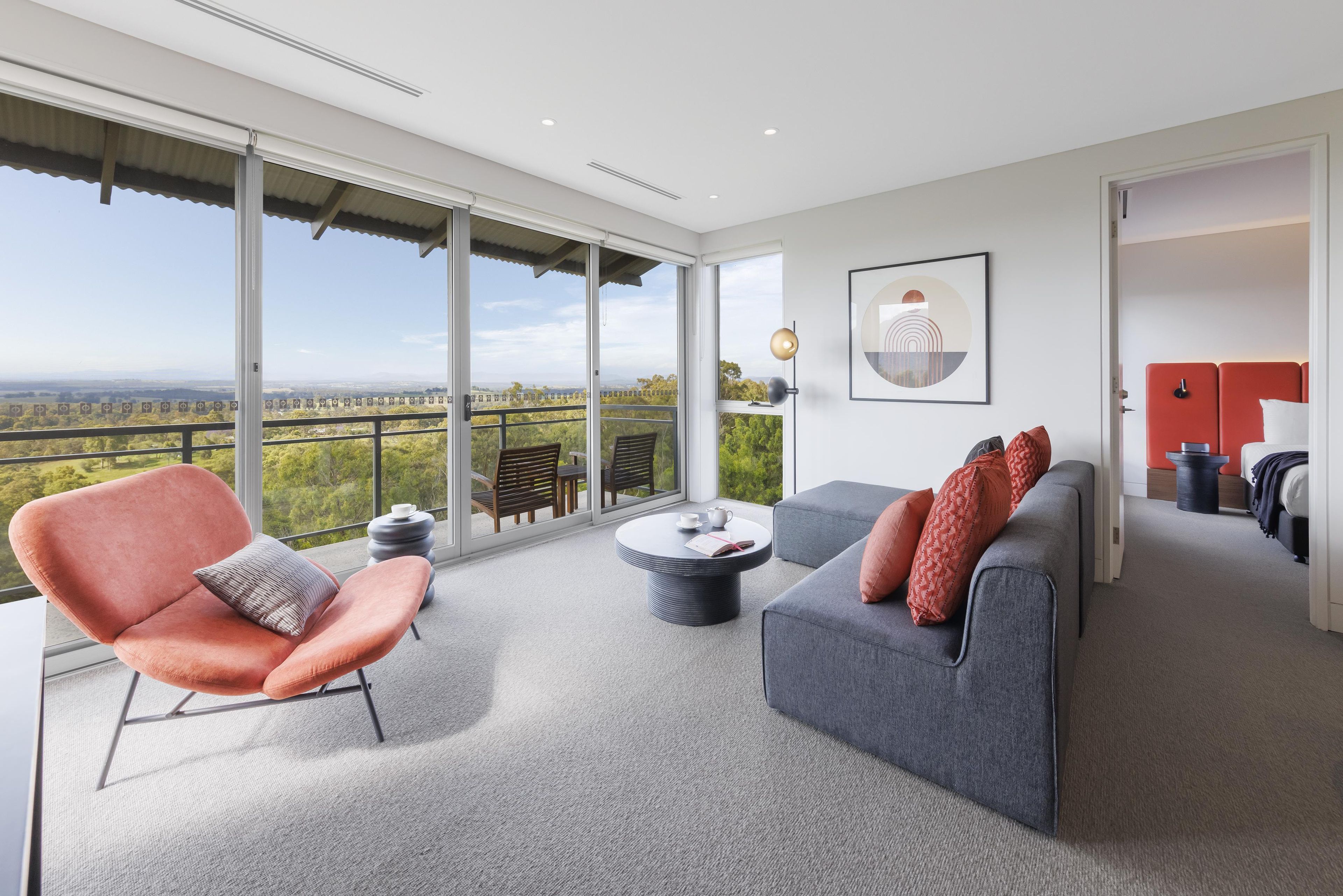 A modern living room with a sofa, armchair, and coffee table, large windows offering a view of the landscape, and a painting on the wall.