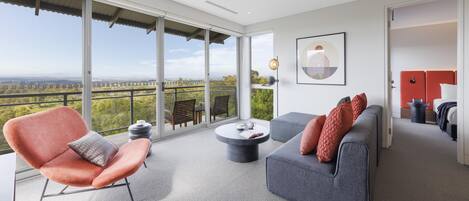 A modern living room with a sofa, armchair, and coffee table, large windows offering a view of the landscape, and a painting on the wall.