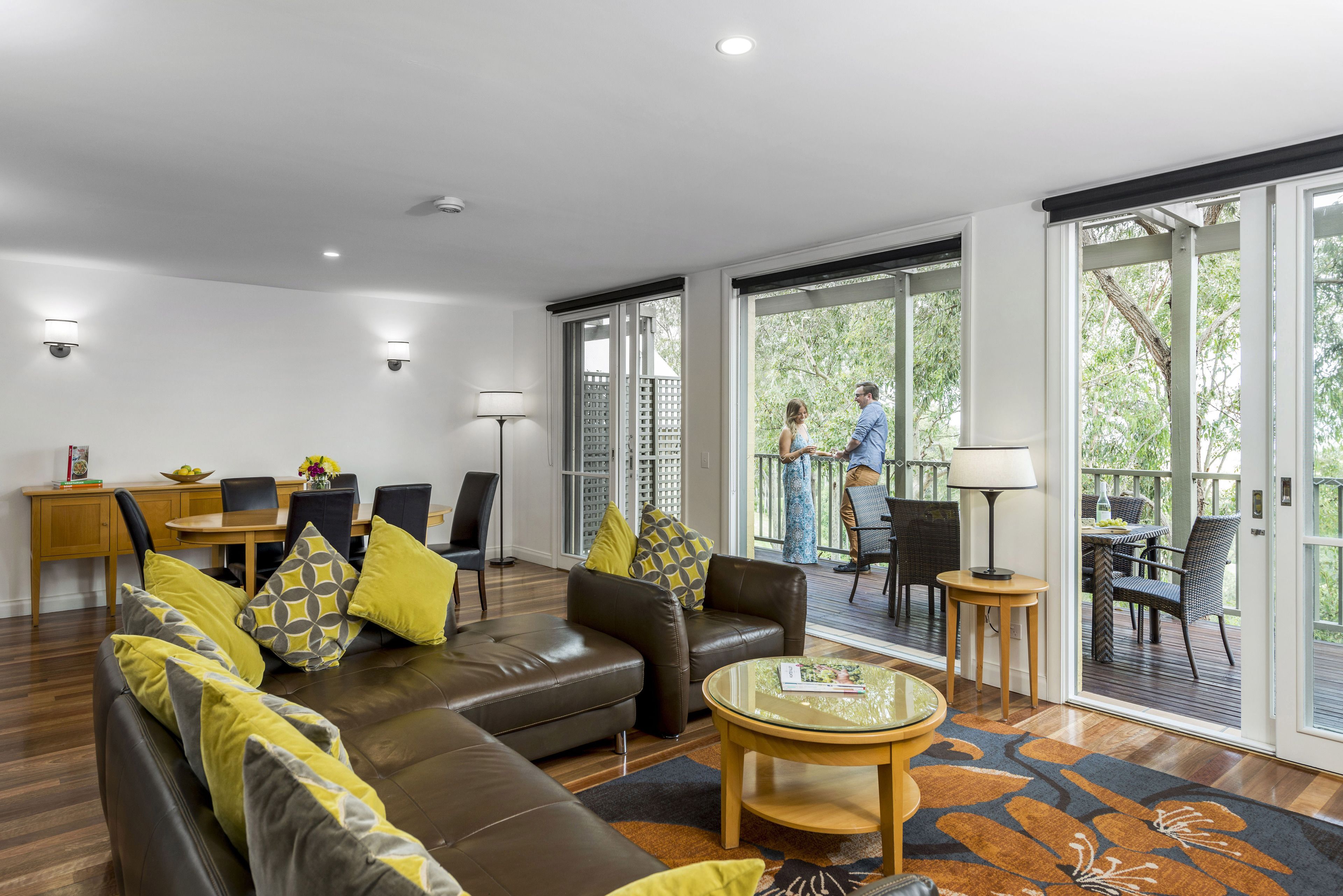Un salon moderne avec un canapé en cuir marron, des coussins aux motifs jaunes et gris, une table basse ronde et un coin repas comprenant une table et des chaises en bois.