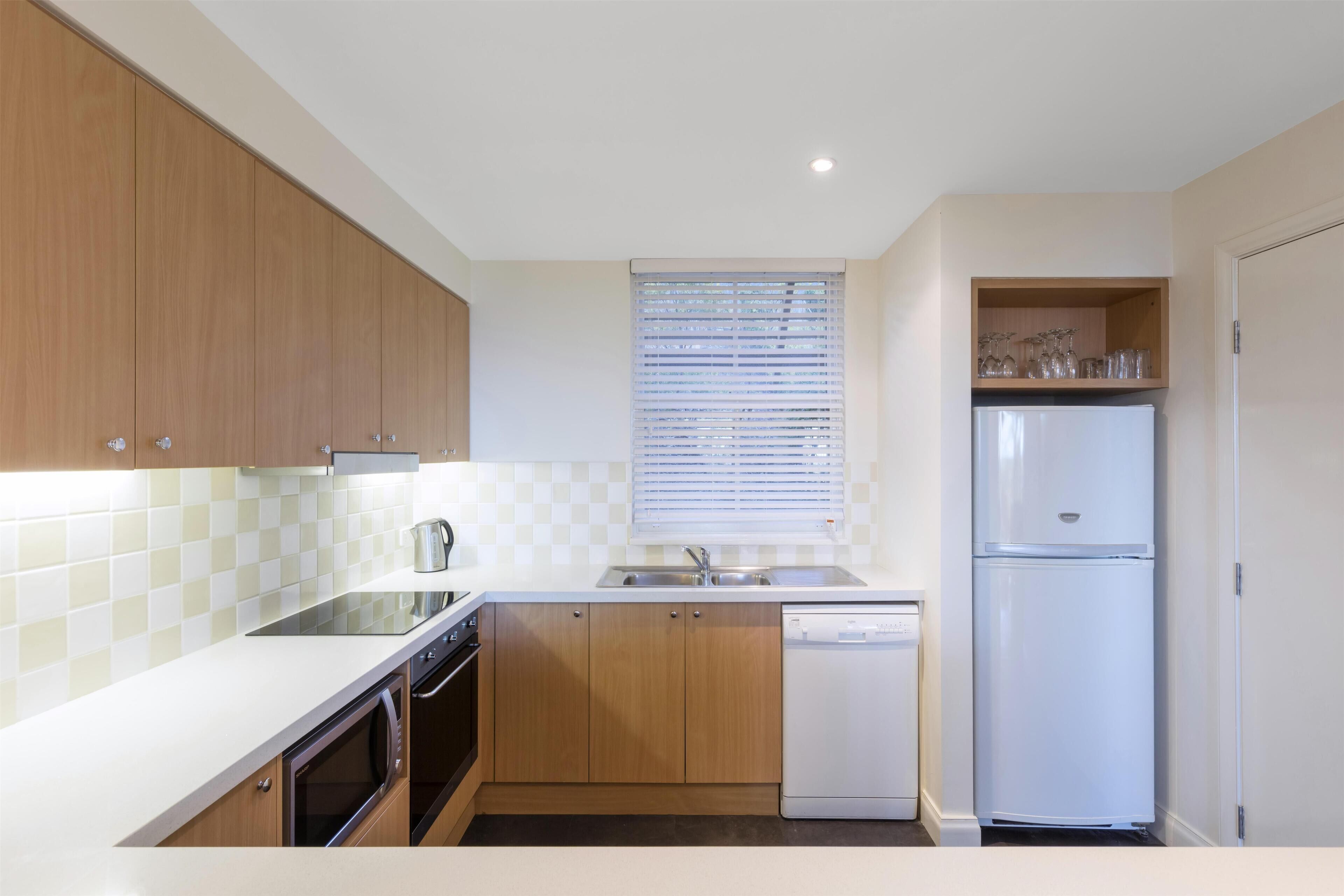 Cocina compacta con armarios de madera, encimera blanca, horno empotrado, lavavajillas y refrigerador blanco.