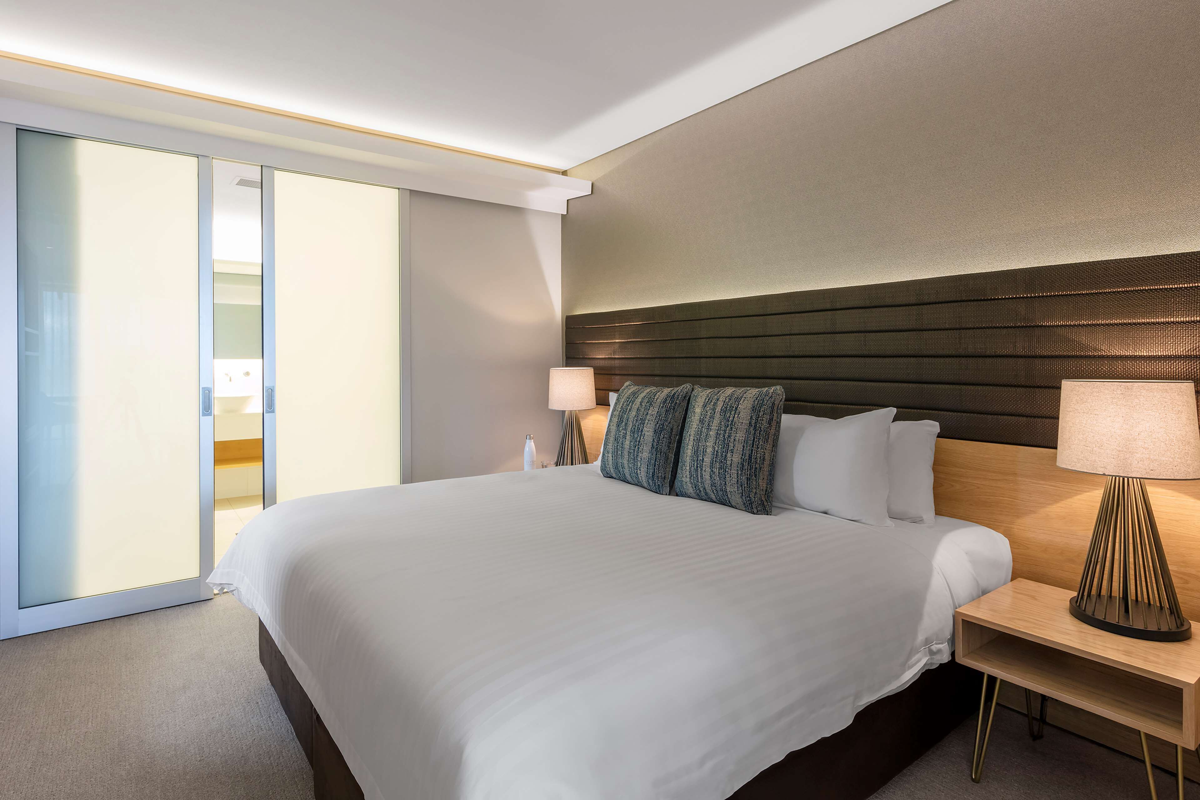 Une chambre d’hôtel avec un grand lit, deux tables de chevet, une lampe et une porte-fenêtre coulissante donnant sur un balcon.