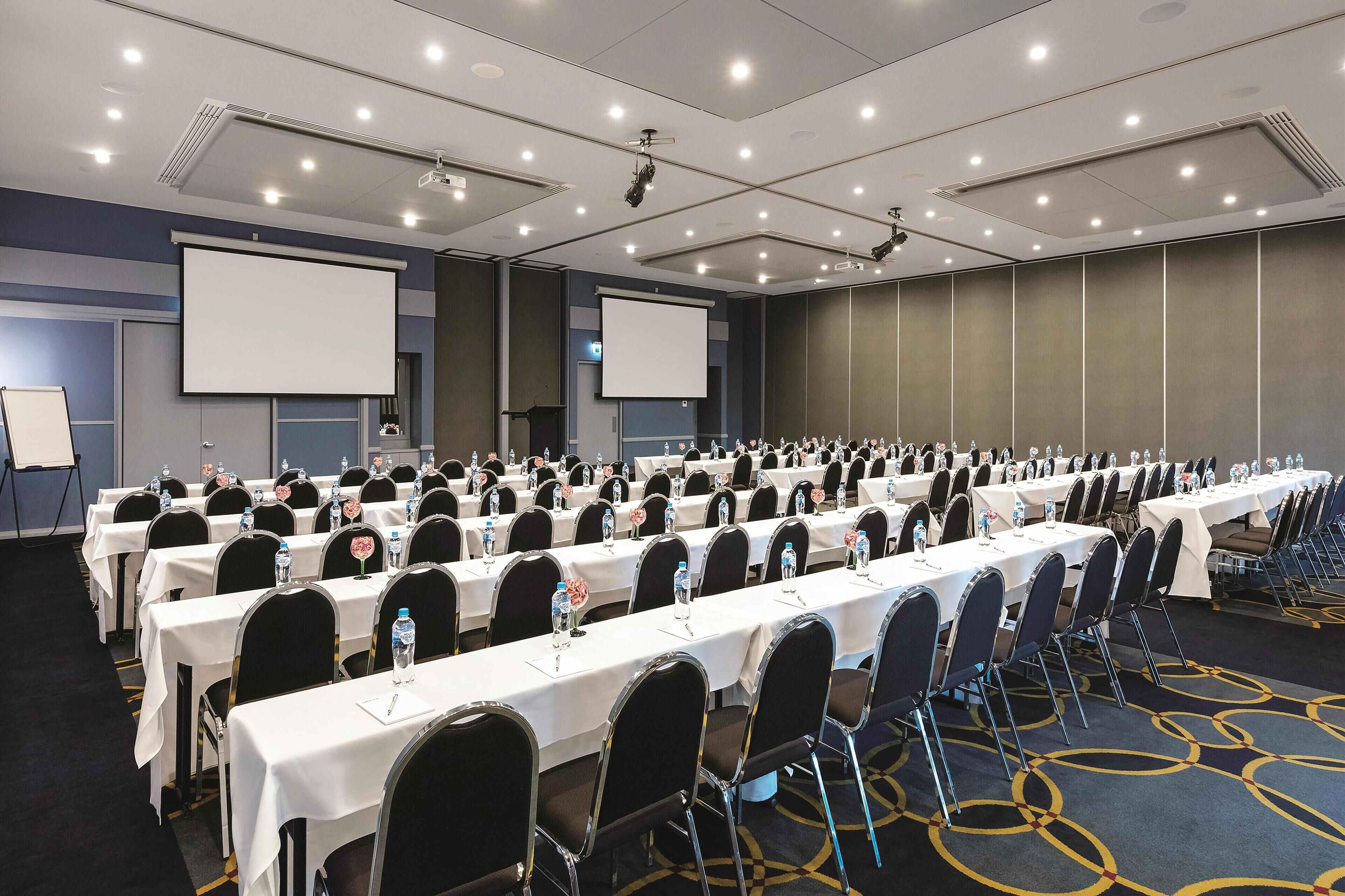 Une salle de conférence avec des rangées de tables, des chaises et un grand écran.