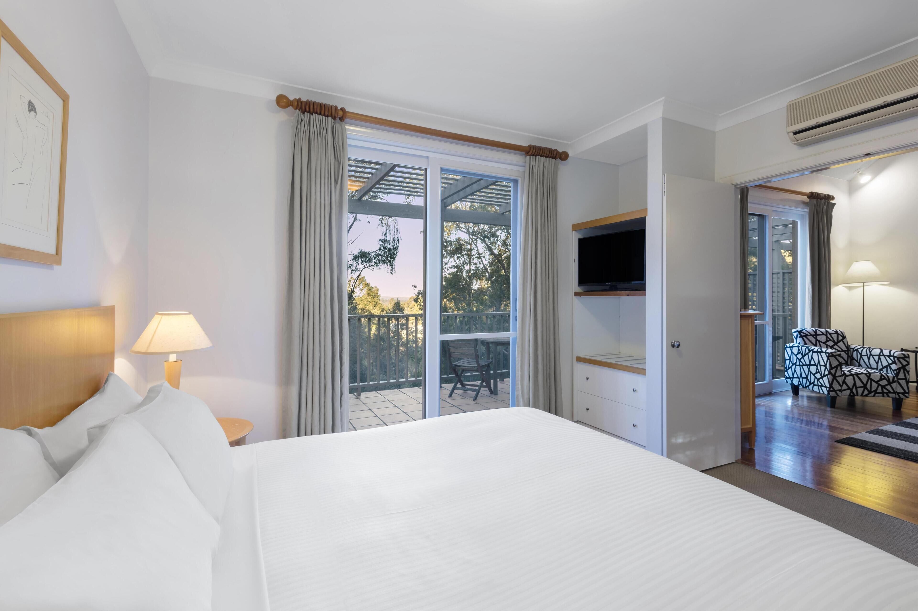 Ein Hotelzimmer mit einem Bett, einem Fernseher, einem Balkon mit einem Sessel und einer Lampe.