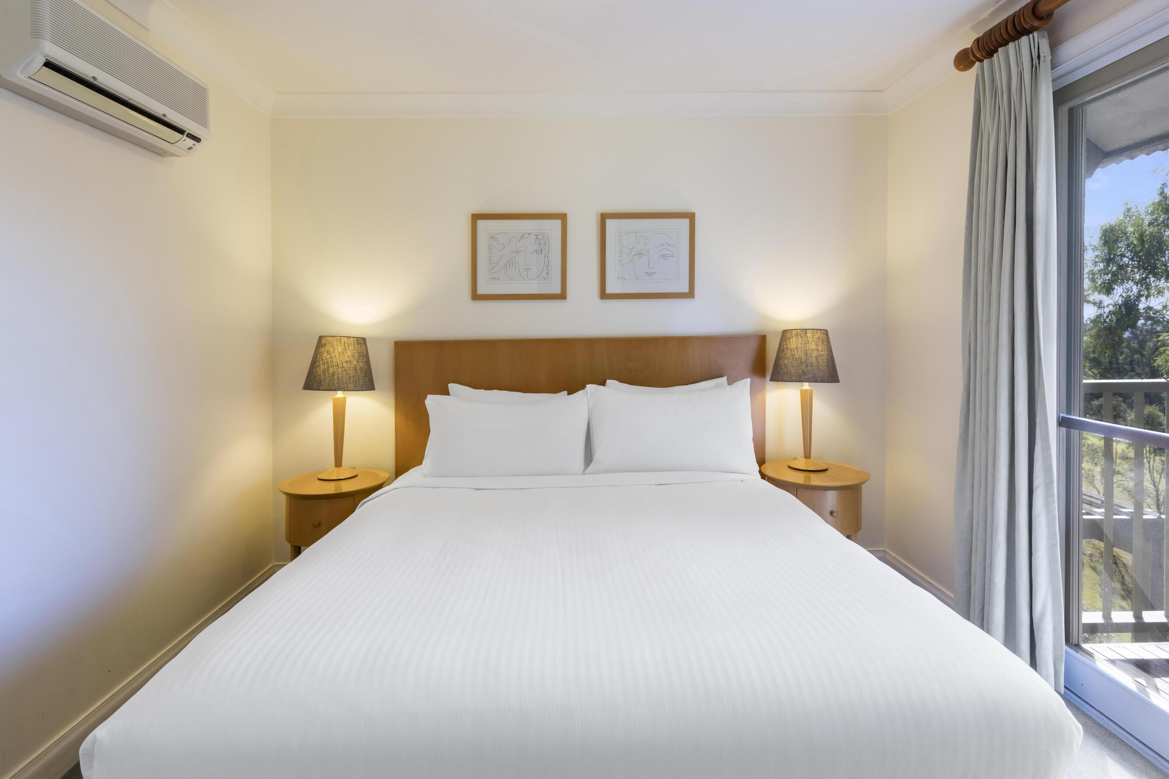Una camera d'albergo con un letto grande, due lampade ai comodini, un condizionatore d'aria e due opere d'arte incorniciate appese al muro.
